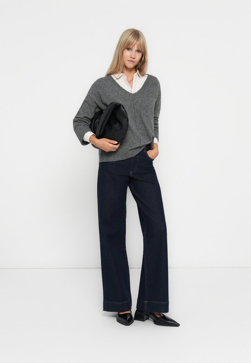 Pull en maille gris à col en V, chemise blanche à col en dessous, jean large noir, et escarpins noirs à bout pointu. Sac noir à la main.