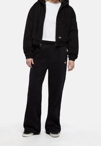 Person wearing black cropped zip-up hoodie, white cropped t-shirt, loose black pants with logo, and black and white sneakers, standing on white background.