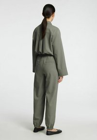 Olijfgroene jumpsuit met een losse pasvorm, lange mouwen en elastische manchetten bij de enkels. Het heeft een ontspannen kraag en een rugplooi detail.