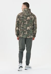 Kamouflagejacka med hög krage, med ett strukturerat mönster i grönt, brunt och beige. Matchad med lätta, mörkgrå joggers.