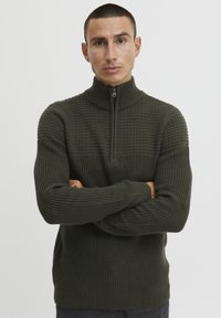 Dunkelgrüner Strickpullover mit hohem Kragen und Reißverschluss, strukturiertem Muster im oberen Bereich sowie gerippten Bündchen und Saum.