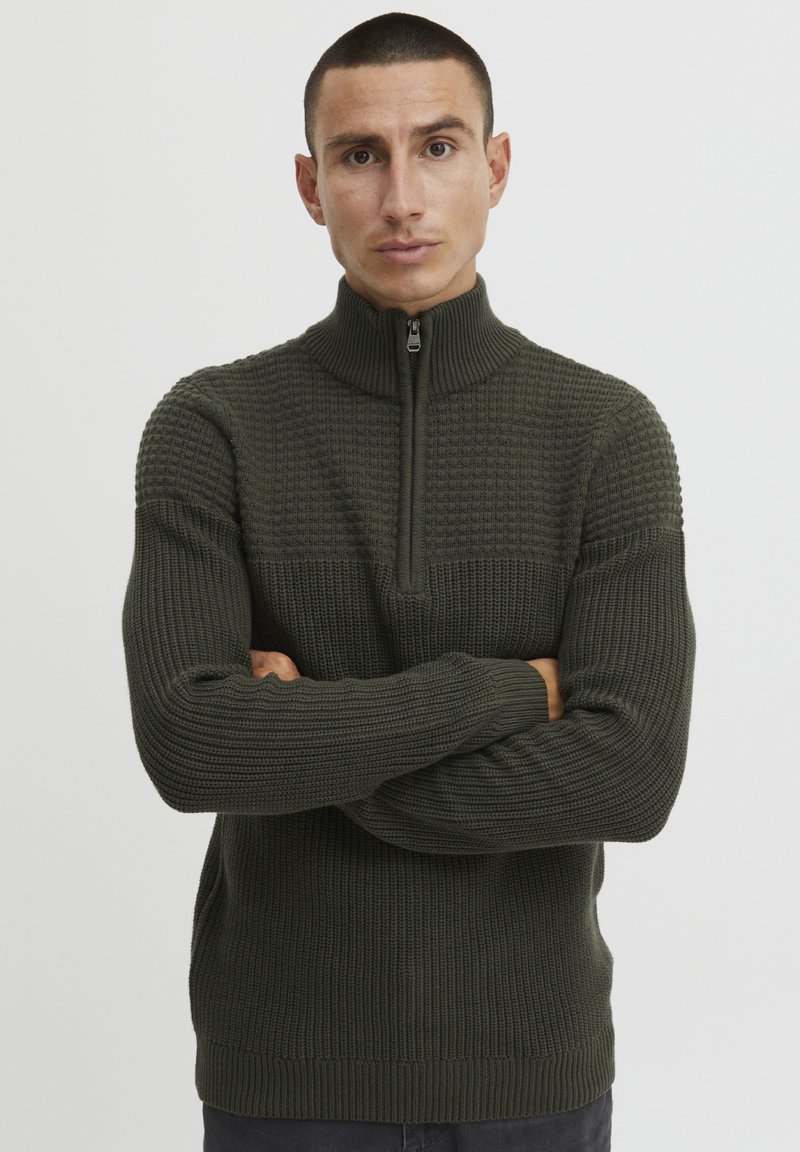 Dunkelgrüner Strickpullover mit hohem Kragen und Reißverschluss, strukturiertem Muster im oberen Bereich sowie gerippten Bündchen und Saum.