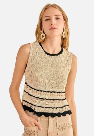 Mujer joven con top de punto sin mangas color beige con borde negro y pendientes, de pie con una mano en el bolsillo contra un fondo blanco.