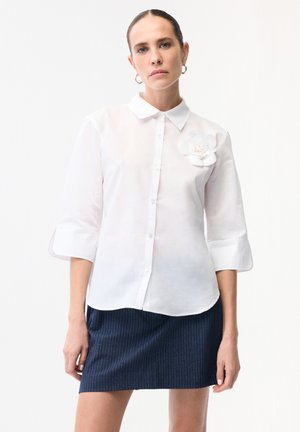 Kobieta z zaczesanymi do tyłu włosami, ubrana w białą koszulę na guziki z rękawami do łokcia i kwiatowym detalem, w połączeniu z ciemną spódnicą w prążki.