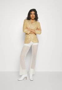 Blazer d'oro con paillettes e colletto a revers abbinato a pantaloni bianchi con motivo in pizzo e stivaletti bianchi con tacco. Texture morbide e dettagli lucidi.
