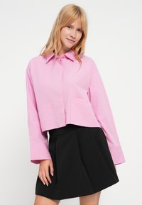 Ροζ κοντομάνικο πουκάμισο με κουμπιά και μακριά μανίκια, με δύο μπροστινές τσέπες, συνδυασμένο με μαύρη φούστα με πιέτες. Λείο ύφασμα και καθαρές γραμμές.