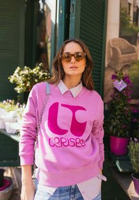 Roze sweatshirt met groot fuchsia-logo, ronde halslijn en lange mouwen, gecombineerd met een lichtblauw overhemd met kraag eronder.