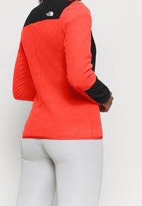 Giacca in fleece di un vivace colore corallo con maniche nere. Presenta un design aderente, una texture morbida e un dettaglio del logo sulla schiena.