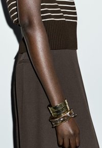 Maglione marrone a coste con strisce bianche, abbinato a una gonna marrone. Tre braccialetti: uno in metallo, uno trasparente e uno in tartaruga. Texture della pelle liscia.