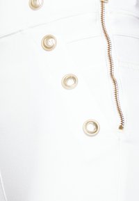 Valkoinen kangas, jolla on sileä rakenne, jossa on kultaisia nappia ja vetoketjun yksityiskohtia. Korostuksia ovat koristeelliset ompeleet saumoissa.