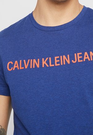 T-shirt en coton bleu à manches courtes avec l'inscription "CALVIN KLEIN JEANS" en lettres orange vives sur la poitrine. Texture lisse.