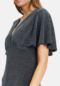 Černé šaty s hlubokým výstřihem do V, s volánovými rukávy a přiléhavým živůtkem z lesklého materiálu, který zdůrazňuje texturu a pohyb.