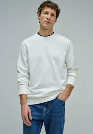 Hombre con cabello rizado que lleva una sudadera blanca de manga larga y jeans azules, de pie con una mano en el bolsillo contra un fondo liso.