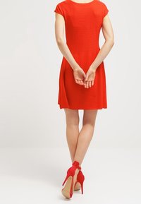 Röd klänning med kort ärm, texturerad kropp och utsvängd kjol. Bärs med röda högklackade skor med en rosett vid vristen.