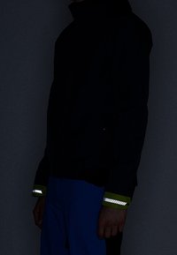 Personne portant une veste sombre avec des bandes réfléchissantes sur les poignets verts et un pantalon bleu sur un fond sombre.