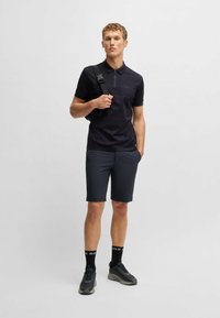 Marineblaues Poloshirt mit kurzen Ärmeln und Reißverschlusshals, versehen mit einem Logodetail, kombiniert mit dunklen Shorts und schwarzen Sportschuhen mit grauen Akzenten.