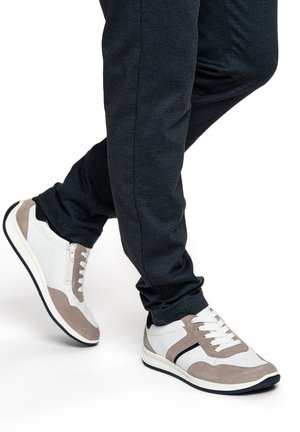 Person trägt weiße und beige Schnürsenkel-Sneaker mit schwarzen Streifendetails und dunkelblaue Hose, steht mit gekreuzten Beinen.
