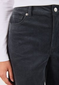 Pantalon en velours côtelé gris foncé avec des côtes verticales, présentant une poche avant, une fermeture à bouton en métal et une texture lisse.