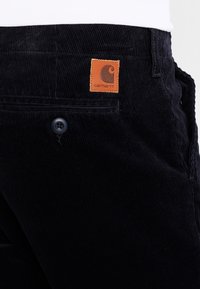 Pantalons en velours côtelé noirs avec une surface texturée, comprenant un patch en cuir marron avec le logo Carhartt, une fermeture à bouton et des poches latérales.