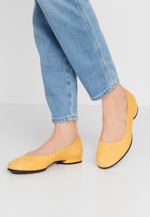 Une personne portant un jean bleu clair et des ballerines en daim jaune avec des talons bas sur un sol gris clair.
