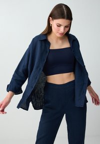 Femme portant une chemise bleu marine ouverte à boutons, un pantalon taille haute assorti et un top tube bleu marine, regardant vers le bas avec les bras légèrement étendus.