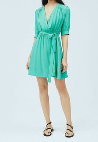 Vihreä kietaisumekko, jossa on lyhyet puffihihat, vyö vyötäröllä ja virtaava helma. Tasainen kangas, rento muotoilu, yhdistetty mustiin sandaaleihin.