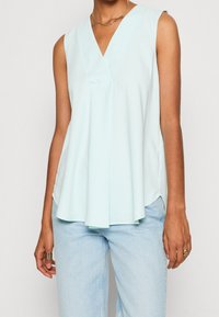 Haut sans manches bleu clair avec un col en V, présentant un tissu drapé et des fentes latérales. Associé à un jean en denim clair. Texture lisse.