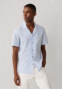 Homme portant une chemise boutonnée à manches courtes bleu clair avec de fines rayures et un pantalon blanc, debout devant un fond uni clair.