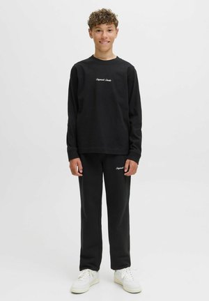 Schwarzer Pullover und passende Hose aus weichem Material mit weißem besticktem Logo auf der Brust und am Bein. Die Sneakers sind cremefarben.