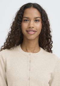 Bež pletena jopica z okroglim ovratnikom in petimi gumbi za zapenjanje, z gladko teksturo in klasičnimi oblikovalskimi elementi.
