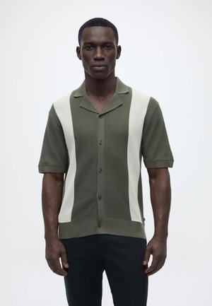 Mann trägt ein olivgrünes, kurzärmeliges Strickshirt mit weißen vertikalen Streifen und steht vor einem schlichten, hellen Hintergrund.