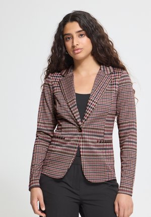 Jeune femme aux longs cheveux bouclés portant un blazer ajusté à carreaux par-dessus un haut noir et un pantalon noir, debout devant un fond uni.