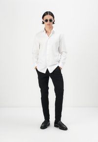 White button-up shirt, black slim-fit pants, black loafers, and round sunglasses. The shirt features a classic collar and long sleeves.