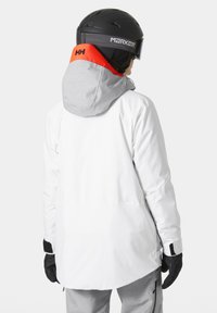 Witte skijas met een grijze en oranje capuchon, met een modern ontwerp, lange snit en handschoenen aan de handen van de drager.