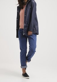 Kvinna i marinblå regnrock över en rosa tröja, uppvikta blå jeans och svarta slip-on skor mot en neutral bakgrund.