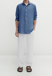 Mann in einem blauen Langarmhemd, weiten weißen Hosen und schwarzen Sandalen vor einem einfarbigen Hintergrund.