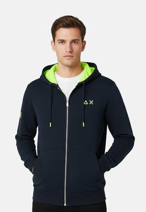 Uomo con felpa nera con cerniera e interno verde neon, piccolo logo geometrico sul petto, mani nelle tasche, maglietta bianca sotto.