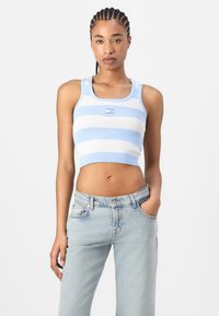 Kort tanktopp med lyseblå og hvite horisontale striper, ribbestruktur og logodetalj på brystet, kombinert med lyse jeans.