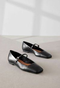 Bailarinas de cuero negro con punta cuadrada, una tira delgada en el empeine y un tacón bajo y plano. Textura suave y diseño minimalista.