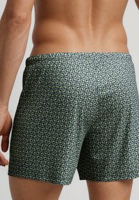 Close-up van de onderrug en bovenbenen van een man in donkere shorts met een herhalend groen, cirkelvormig geometrisch patroon.