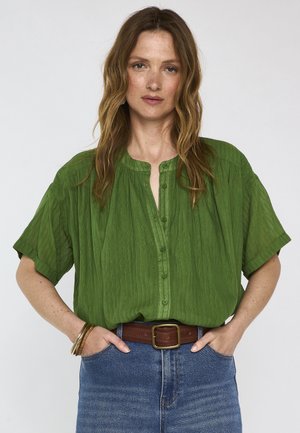 Vrouw met golvend bruin haar die een losse groene blouse met knopen draagt, blauwe jeans, een bruine riem en gouden armbanden, met haar handen in de zakken.
