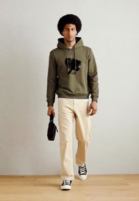 Sweat à capuche vert olive avec motif graphique noir, pantalon crème et baskets noires. Portant un petit sac noir, debout sur un sol en bois.