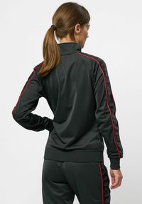 Schwarze Sportjacke mit hohem Kragen, ausgestattet mit roten Seitenstreifen und strukturierten Einsätzen. Glattes Material und taillierte Passform.
