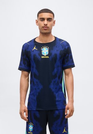 Giovane uomo con capelli corti che indossa una maglia da calcio e pantaloncini blu scuro del Brasile con dettagli dorati e verdi, in piedi davanti a uno sfondo semplice.