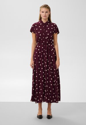 Femme portant une robe midi bordeaux à pois blancs, manches courtes, col et ceinture nouée à la taille, debout sur un fond clair.