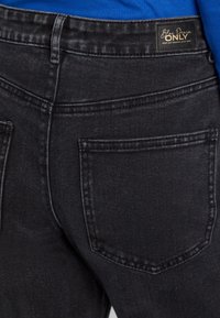 Černé džíny z denimu s detailním pohledem na zadní kapsy, jemnou texturu a štítek s nápisem "Blue Denim ONLY" na pásku.