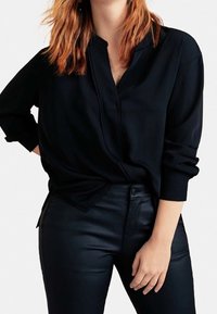 Schwarze Bluse mit langen Ärmeln, Stehkragen und dezenten Falten; kombiniert mit eng anliegenden schwarzen Hosen. Glattes Material, lockere Passform.