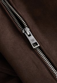 Close-up of a metal zipper on dark brown suede fabric, with "ESTD 1982" engraved on the zipper pull.