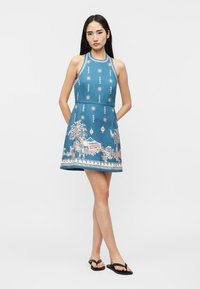 Γυναίκα με αμάνικο μπλε φόρεμα με στάμπες ήλιου και φοίνικα, στέκεται με τα χέρια πίσω από την πλάτη, φοράει μαύρα σαγιονάρες, ουδέτερο φόντο.