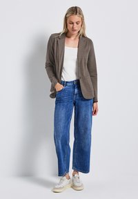 Blazer long gris à manches longues sur un top blanc, associé à un jean en denim bleu taille haute. La tenue est complétée par des baskets blanches.